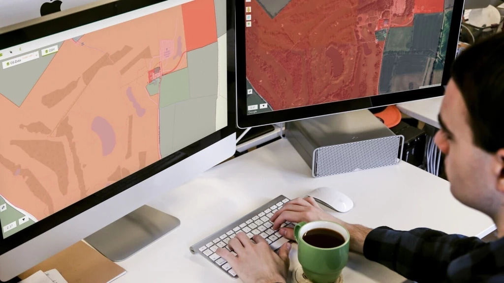 Planner working at a desk with two monitors displaying detailed land-use and Green Belt maps, analysing development and economic impacts.