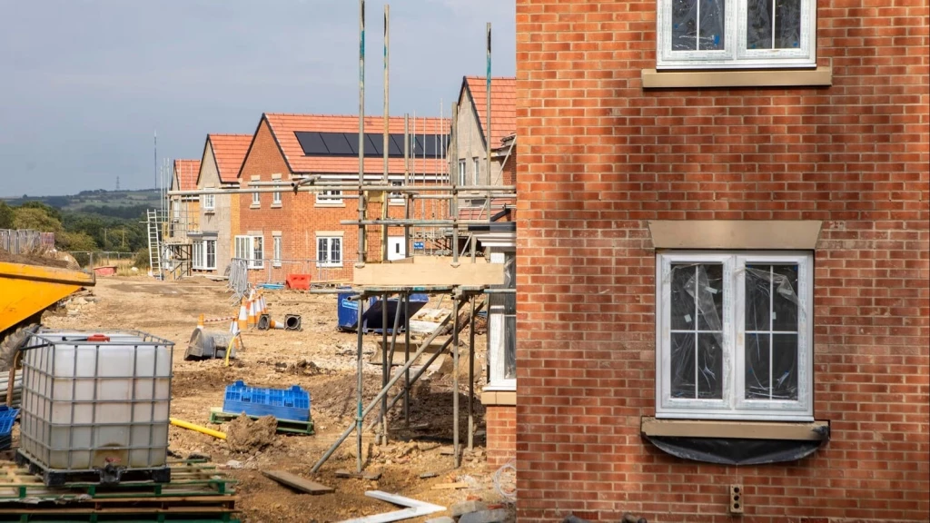 New-build houses under construction with scaffolding, site machinery and solar panels, highlighting development profits created by restricted land supply.