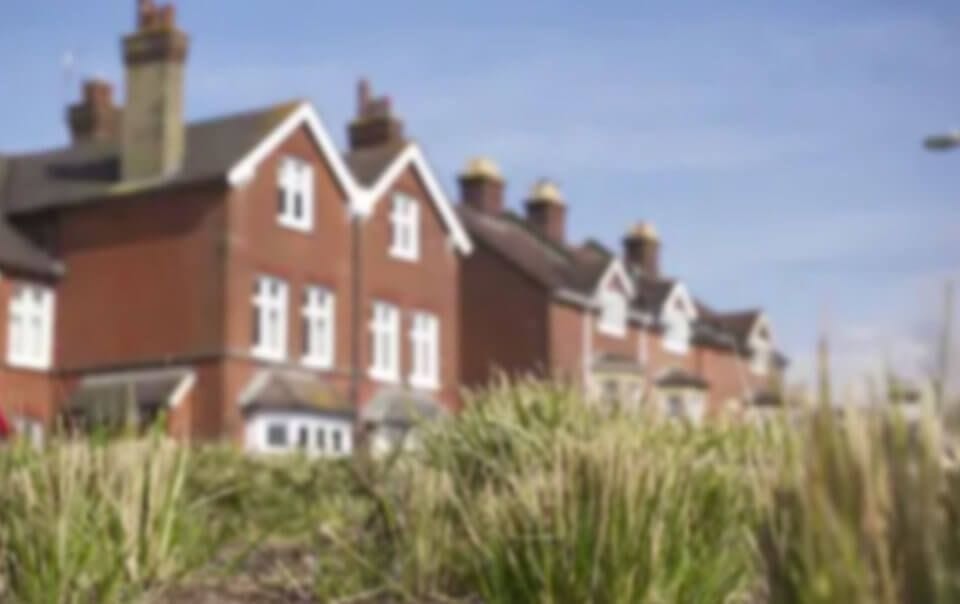 Unfocused photograph taken from between green grass blades facing three storey traditional red brick and white trimmings detached homes with clear blue skies