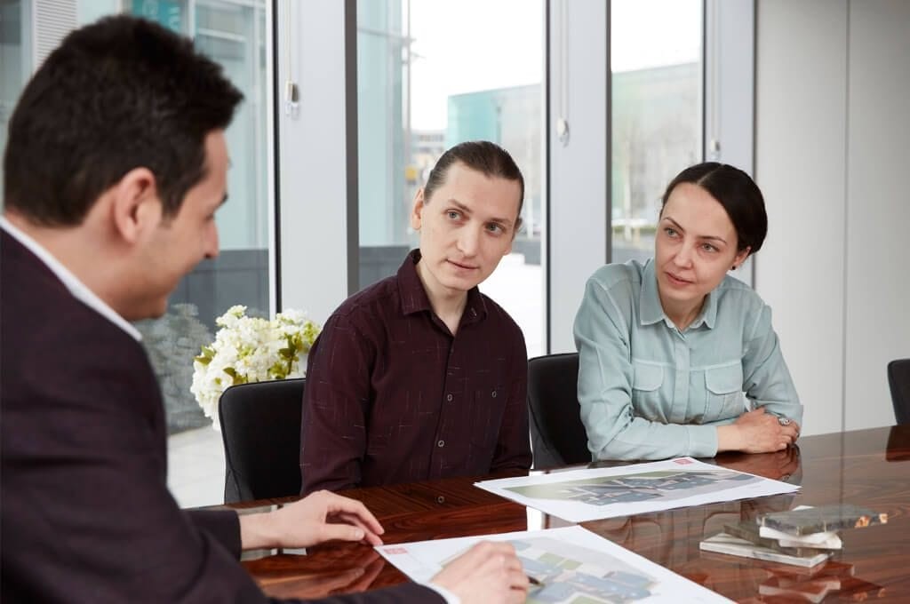 Three professionals in a focused meeting at a modern office. One man, facing the camera, discusses architectural plans spread on the table, while a male and a female colleague attentively listen. The background features a glass facade, providing a view of the exterior urban setting. A vase of white flowers adds a touch of elegance to the meeting environment.