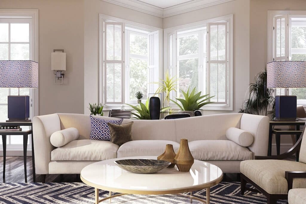Elegant and serene living room in a London home with large bay windows providing natural light, showcasing two plush cream sofas with decorative cushions, a round white coffee table, patterned lamps, and indoor plants, reflecting a classic yet modern interior design aesthetic.