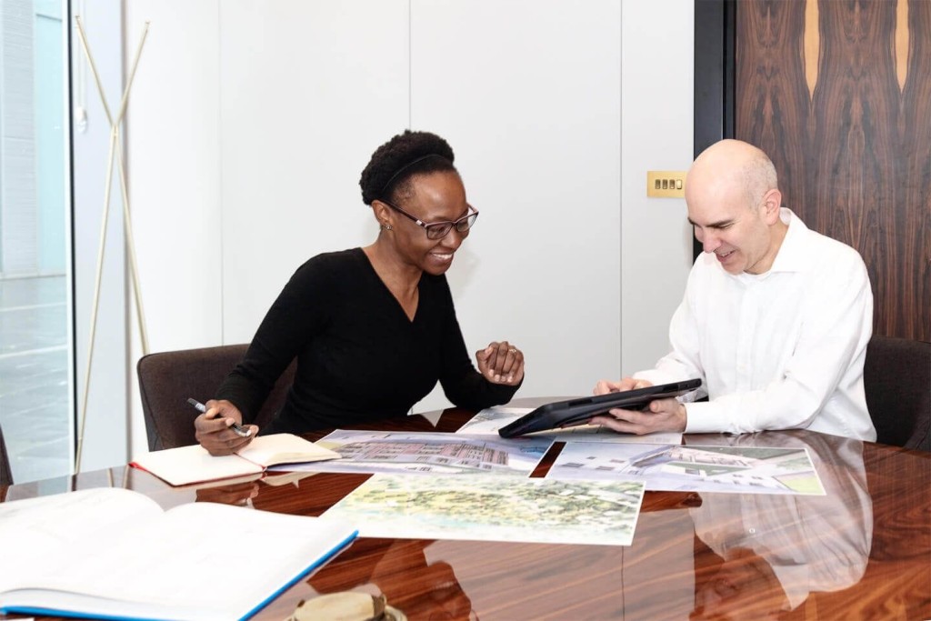 Two architects in a professional meeting, reviewing project plans on a digital tablet, with building blueprints and design illustrations spread on a wooden table in a modern office environment.