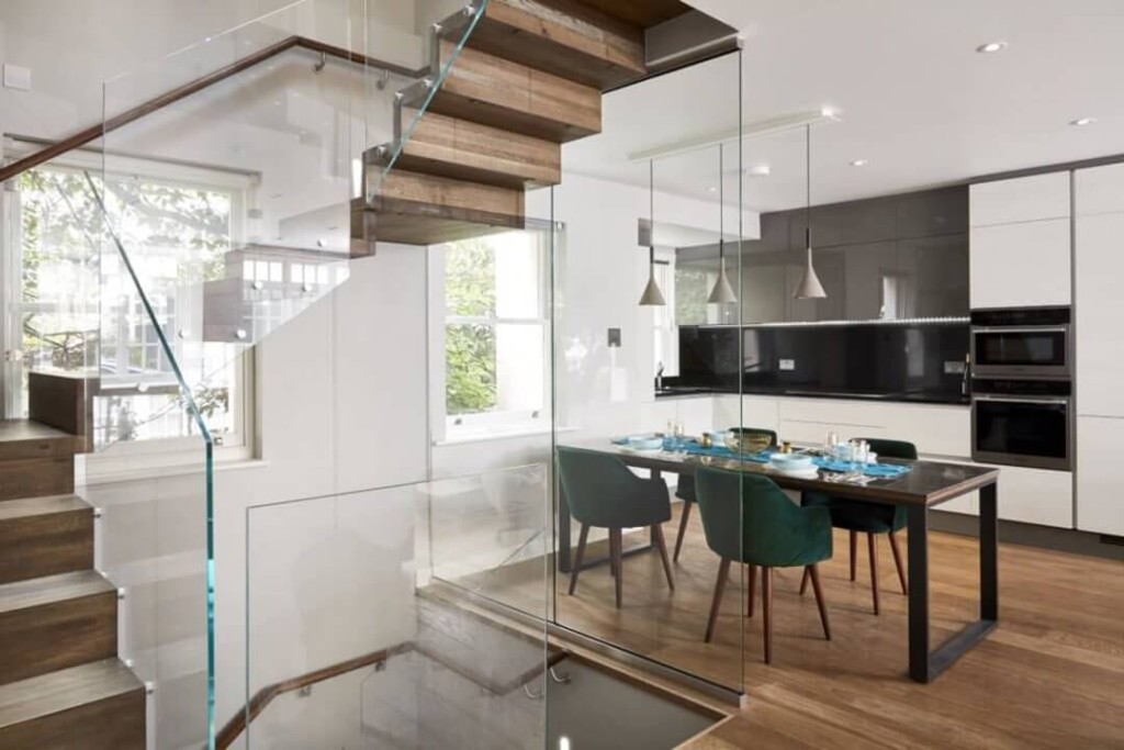 Modern London home interior with an open-plan kitchen and dining area; features include sleek white cabinetry, dark countertops, high-end appliances, a wooden staircase with glass balustrade, and a dining table set with forest green chairs, showcasing contemporary design and luxury living.