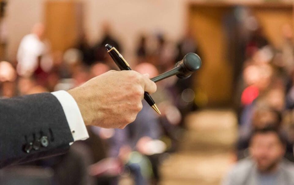 Close up of a male arm and hand in a two piece dark grey suit holder a black and gold pen as well as black gavel extending towards the blurred audience at a auction sale