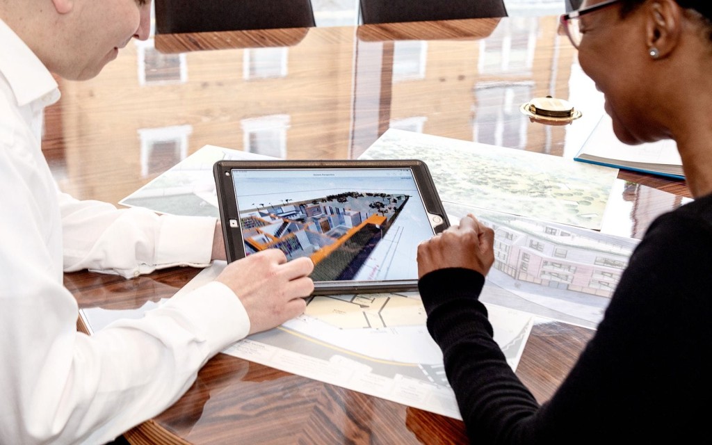 Two architects collaborating over a digital tablet displaying a 3D architectural model, with building plans spread out on a glossy wooden table, in a bright office setting with reflections on the surface indicating a creative work environment.