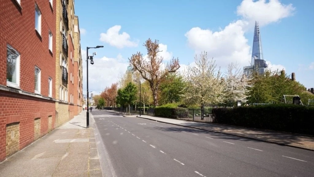 Empty urban street in a sunny day with blossoming trees lining the sidewalk, red brick apartment building on the left, and the iconic Shard skyscraper visible in the background against a clear blue sky in London.