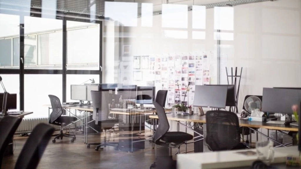 Modern open-plan office space bathed in natural light, featuring multiple workstations with computer monitors, rolling chairs, and a collaborative atmosphere, reflecting contemporary corporate interior design.