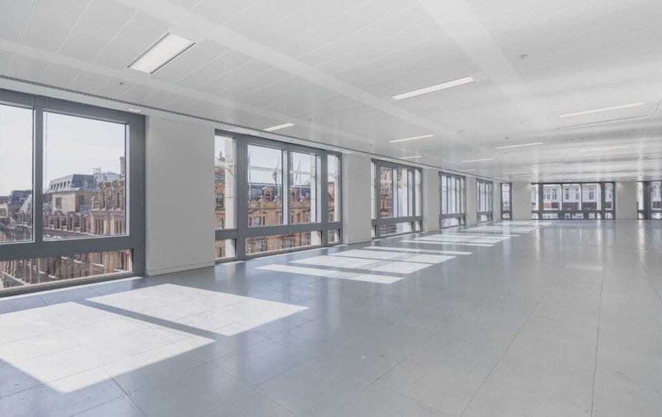 Spacious and empty office interior flooded with natural light, showcasing a modern open-plan space with a series of large windows offering a panoramic view of the city, illustrating potential for conversion into residential living spaces.