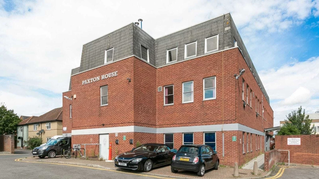 Paxton House in London, a modern red brick building on a sunny day, showcasing its angular architecture and large windows, with clear signage on the facade, surrounded by parked cars and urban landscaping, reflecting contemporary city living and office space design.