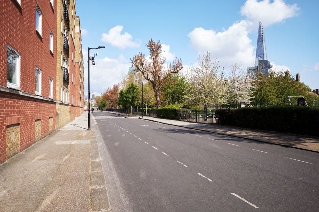 Empty urban street on a sunny day with blossoming trees lining the sidewalk, red brick apartment building on the left, and the iconic Shard skyscraper visible in the background against a clear blue sky in London.