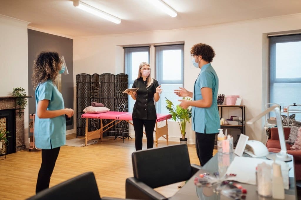 Nail technicians and aesthetician standing in the middle of the salon with protective masks and face shields in an impromptu meeting