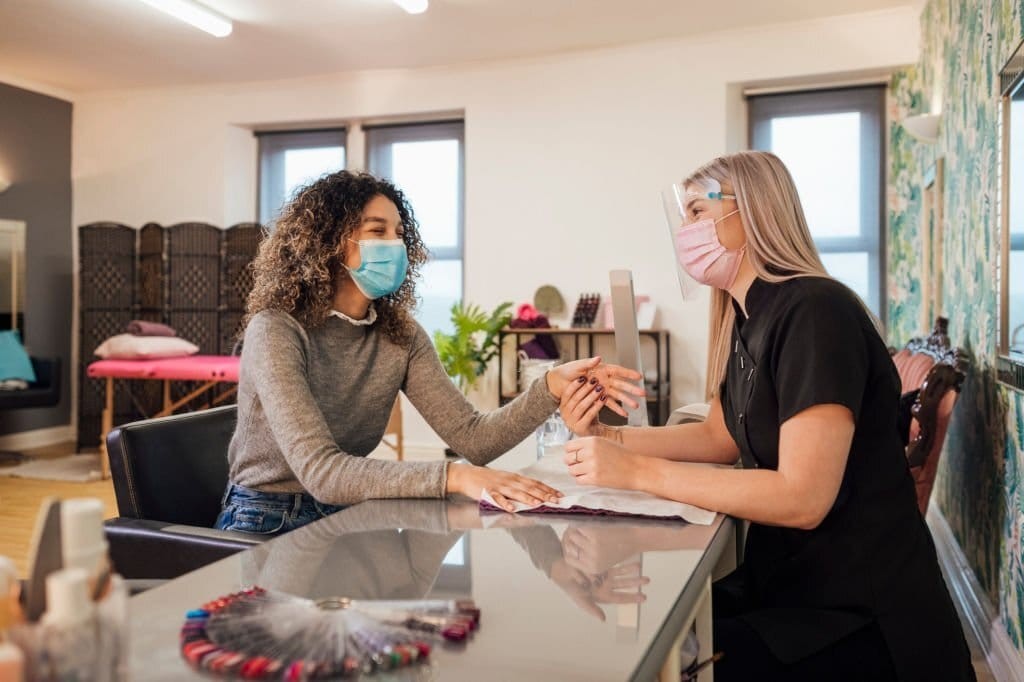 Client and nail technician laughing behind their protective covid-19 masks whilst having a manicure done and a splay of nail swatches is left on the table next to them