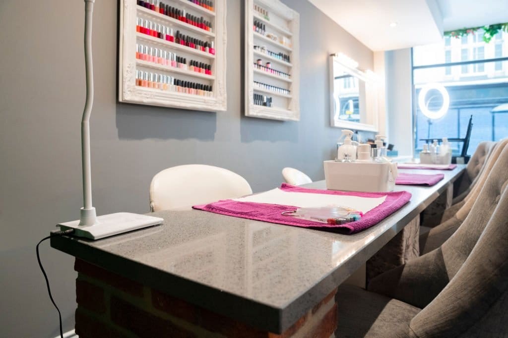 Modern yet simplistic interior design of a nail salon with grey walls, table tops and plush client chairs and white lights and white nail polish display cases.