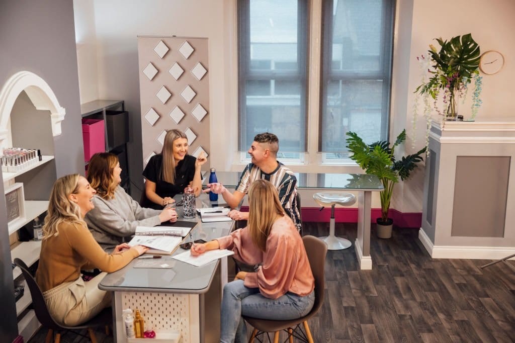 Aesthiticians, hairdressers and nail technicians all gathered around a grey table on the side of the salon for a smiling team meeting