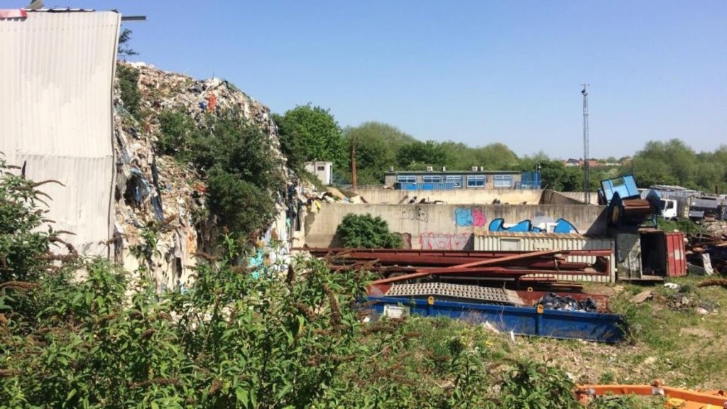 Overgrown and cluttered brownfield site with heaps of industrial waste and scattered construction materials, including a large mound of debris, hinting at the potential for redevelopment and the challenges of environmental management within UK Green Belt areas.