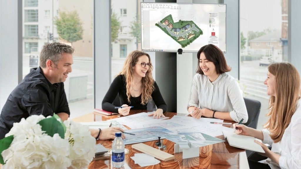 Four urban planners in a bright office environment engaged in a collaborative discussion over architectural blueprints, with a digital 3D model of a Green Belt development project displayed on a screen in the background, exemplifying a professional consultation for sustainable urban planning.