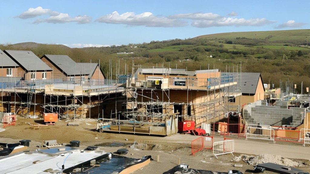 New housing development on brownfield land with partially constructed homes and scaffolding, highlighting ongoing construction work amidst a rural backdrop with rolling hills, under a clear sky, emphasising the complexity of solving the UK's housing shortage with brownfield sites alone.