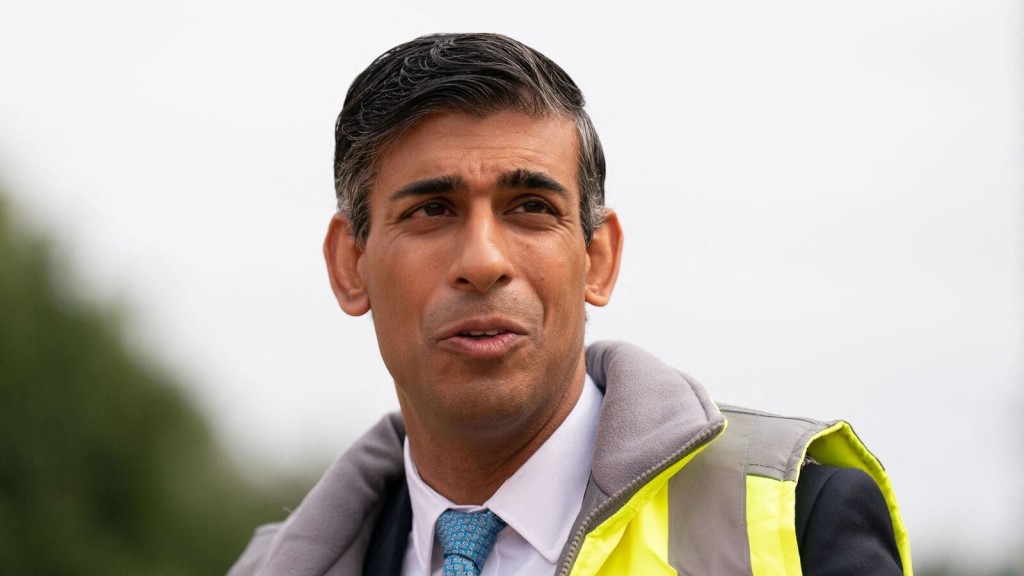 Rishi Sunak with a high-visibility vest stands outdoors, with a concerned expression possibly addressing urban development issues, representing the involvement of policymakers in decisions regarding construction on Green Belt land.