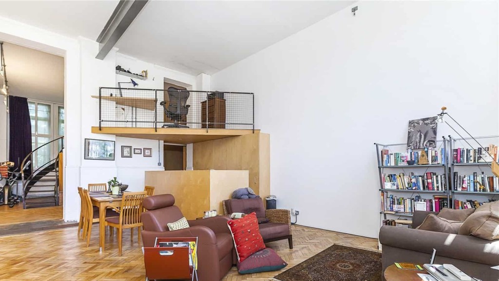 Spacious living area in a listed building featuring high ceilings, a cosy reading nook, herringbone wood flooring, and a simple mezzanine level with a metal railing.