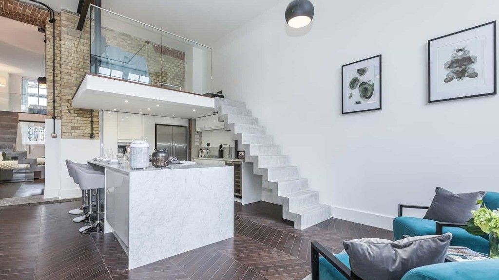Modern and stylish maisonette in a listed building featuring exposed brick walls, sleek white kitchen island with bar stools, a floating staircase, and a comfortable living area with teal arm chairs. 
