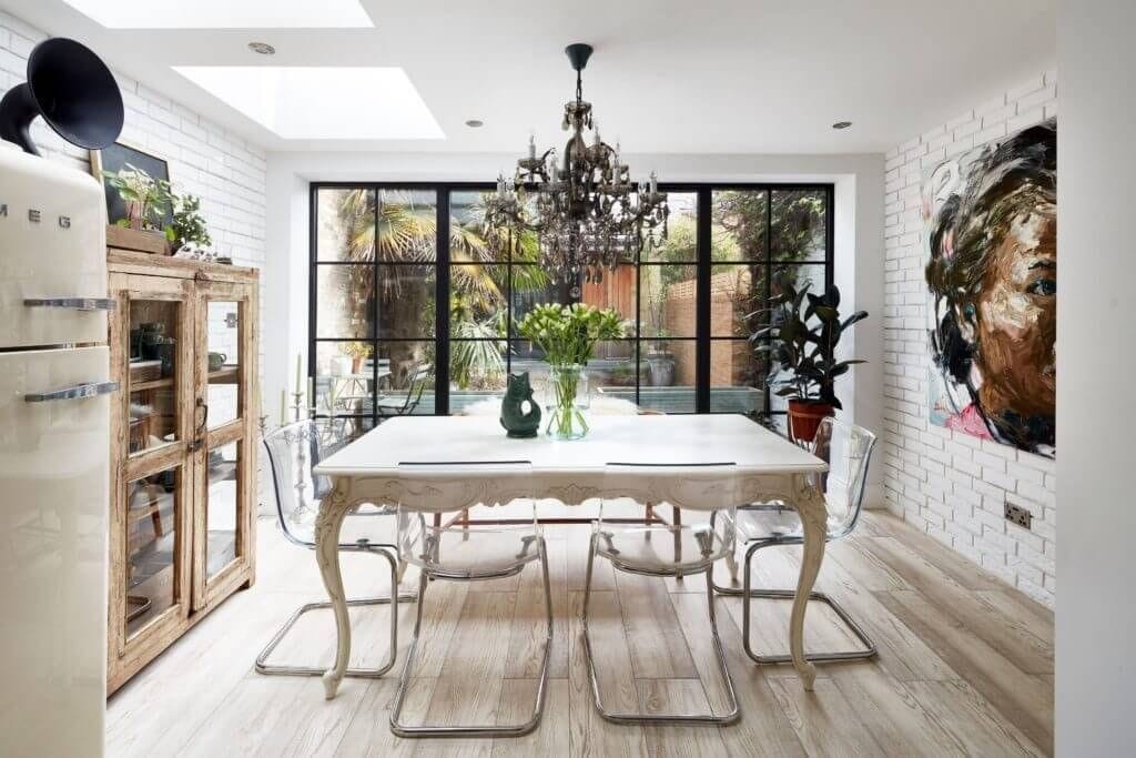 Beautiful kitchen extension in a London home featuring a blend of rustic and contemporary design elements. The space includes a vintage-style dining table with clear acrylic chairs, a statement chandelier, and large glass doors that open to a garden. White brick walls and a retro SMEG refrigerator enhance the charm, while a large modern painting adds a touch of artistic flair. Perfect for homeowners looking to create a stylish and functional kitchen extension with an eclectic mix of old and new.