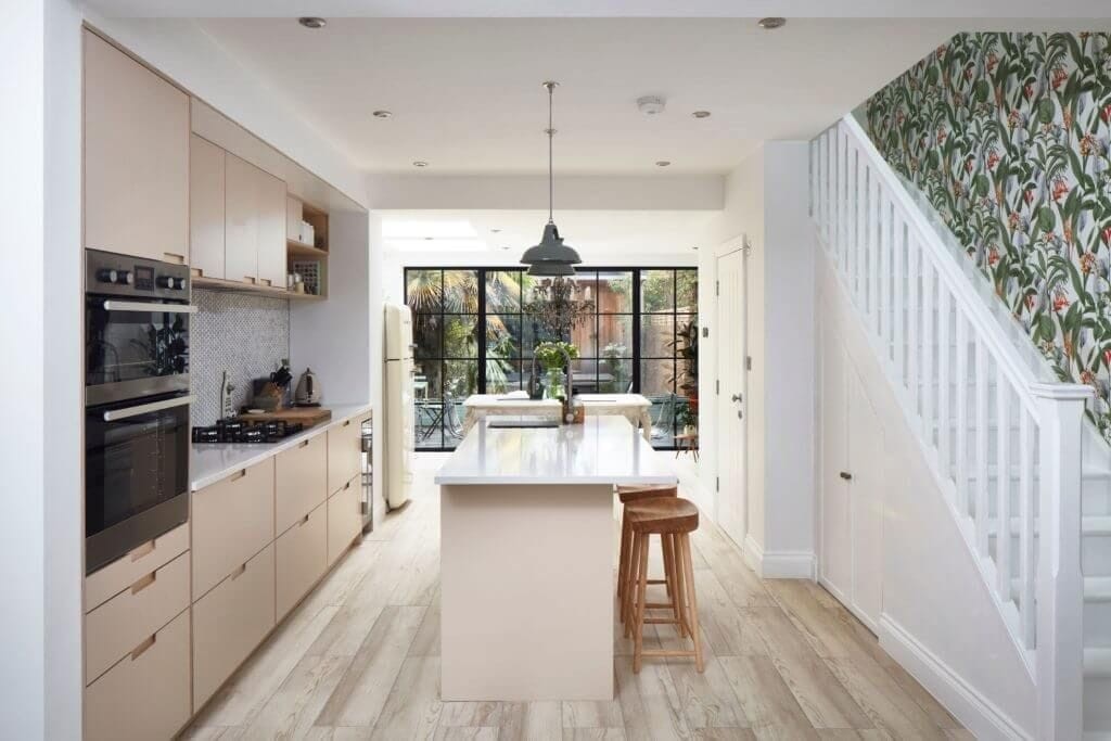Charming kitchen extension in a London home featuring pastel cabinetry, a central island with bar stools, and modern appliances. The design includes a large glass door that opens to a lush garden, maximising natural light. A unique floral wallpaper accents the staircase, adding a touch of personality to the bright, airy space. Ideal for homeowners seeking a blend of contemporary and cosy elements in their kitchen renovation.
