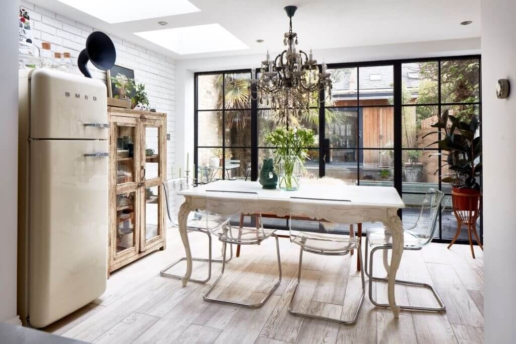 Elegant kitchen extension in a London home featuring a mix of rustic and modern design elements. The space includes a vintage-style dining table with clear acrylic chairs, a statement chandelier, and large black-framed glass doors that open to a garden. The kitchen also boasts a retro cream SMEG refrigerator and a rustic wooden cabinet. White brick walls and skylights enhance the natural light, creating a bright and inviting atmosphere. Ideal for homeowners seeking a stylish and functional kitchen extension with a blend of old-world charm and contemporary flair.