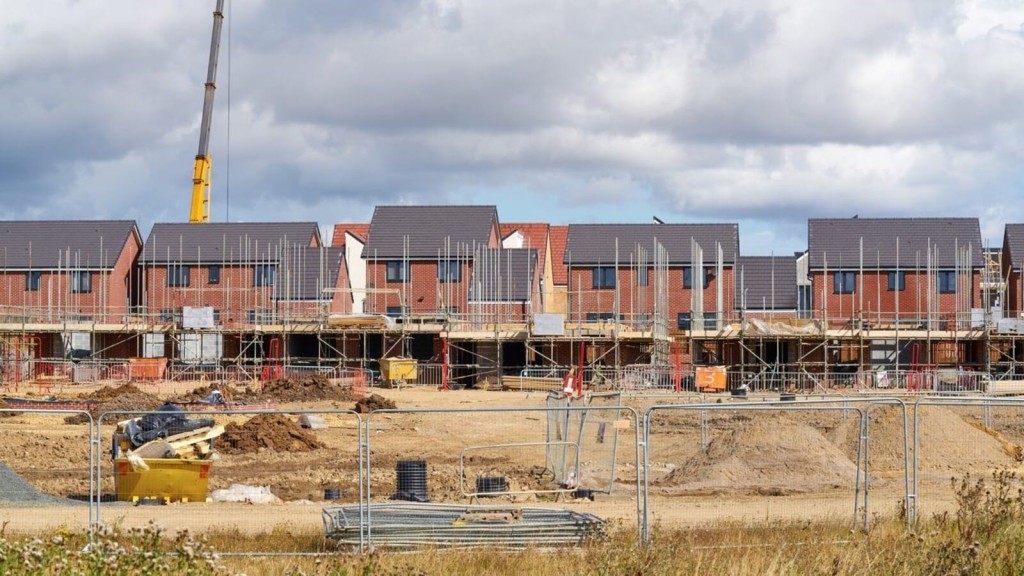 New residential development under construction with row of semi-detached houses, scaffolding, and construction crane on a clear day, depicting ongoing housing projects in the UK.