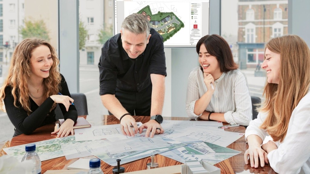 Team at Urbanist Architecture consisting of architects and planners engaged in a collaborative meeting around a wooden table, discussing a residential building project. A digital screen in the background displays a site plan. The group, consisting of one man and three women, are smiling and actively participating, highlighting a productive and innovative work environment.