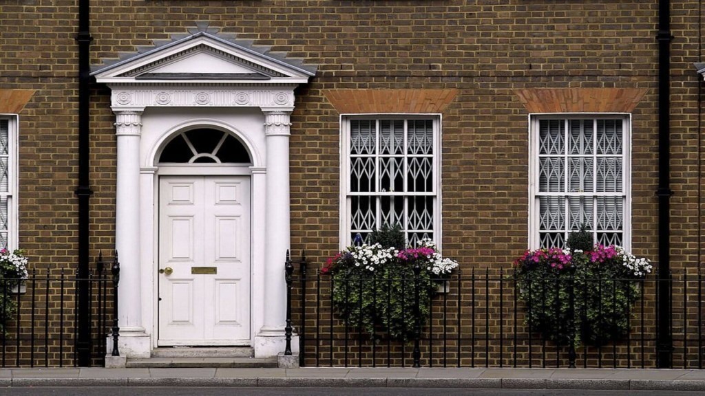 Traditional Georgian style facade with a white front door and ornate portico, symmetrical sash windows with decorative iron bars, and blooming flower boxes, set against a classic brick wall on a historical street.