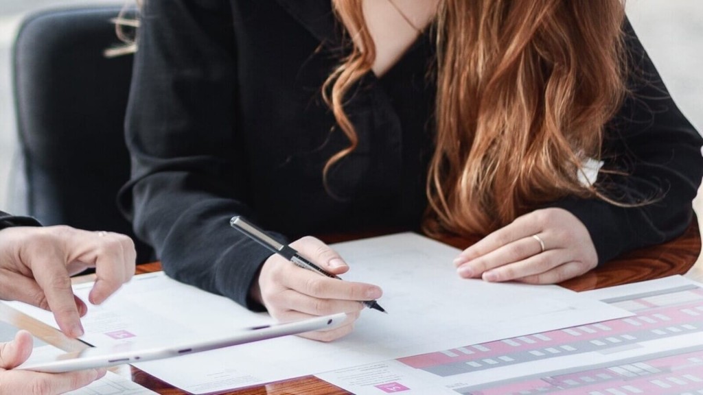 A close-up of two individuals collaborating over a set of architectural plans, focusing on a woman's hands holding a pen, poised to make notes, with detailed building schematics visible on paper, implying a professional and concentrated work environment.