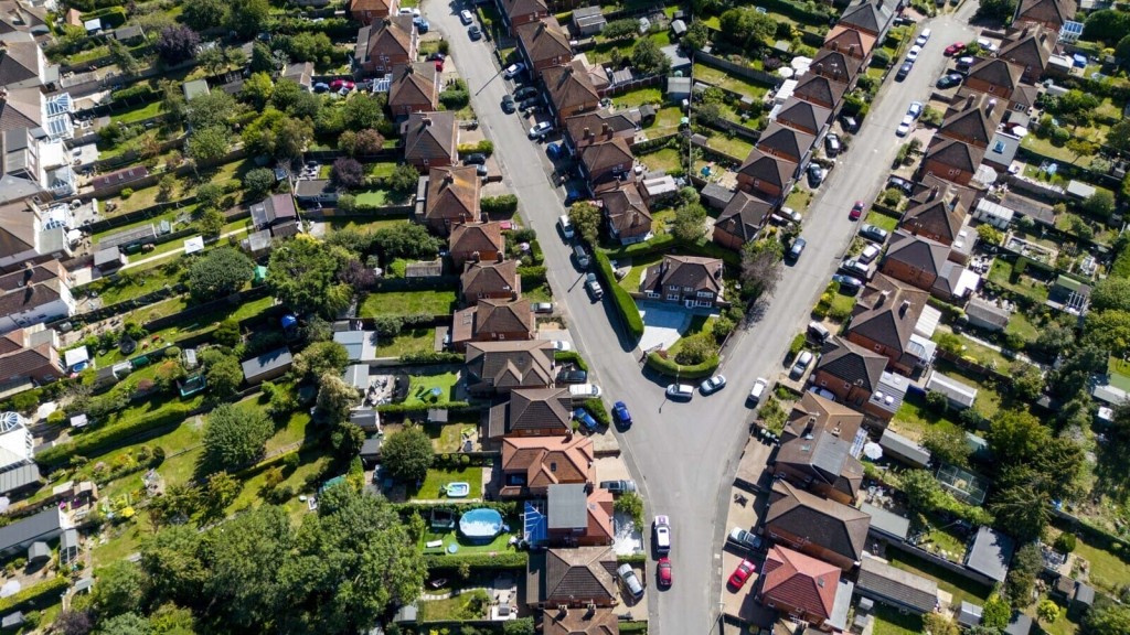 Aerial view of a suburban neighborhood showing a crossroad, residential houses with pitched roofs, neat backyards, and lush greenery on a bright sunny day, depicting peaceful community living.