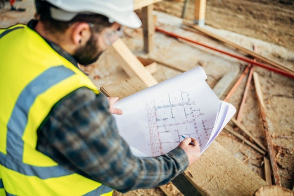 Construction professional in high-visibility vest and hard hat reviewing blueprints on a building site, with focus on detailed architectural plans and wooden framework in the background.