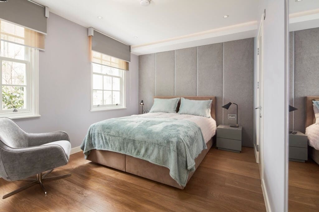 Inviting bedroom featuring a plush bed with a soft teal bedspread, padded grey headboard, hardwood flooring, and a cosy reading nook with a stylish grey armchair, lit by natural light from two sash windows.