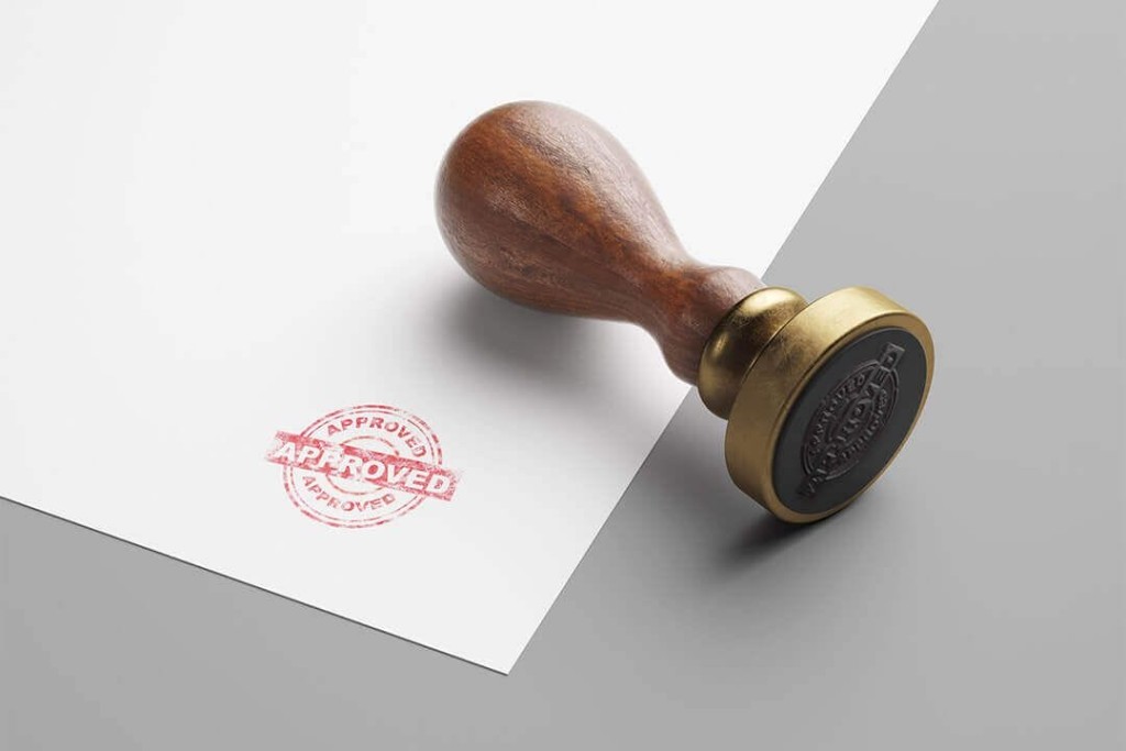 Classic wooden and brass seal stamper with 'APPROVED' stamp on white paper, symbolising official approval, with a sharp shadow on a light grey background, representing authority and authentication.