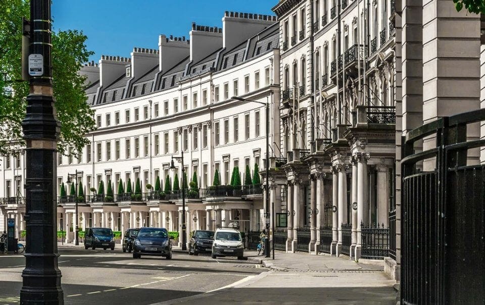 Elegant Georgian townhouses line a peaceful street in Belgravia, London, featuring classic white facades and columned entrances, with greenery and parked cars under a clear blue sky.