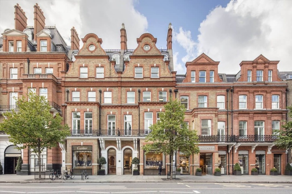 Victorian red brick buildings with ornate gables and wrought iron balconies on a London street, featuring ground floor luxury retail stores including Cartier, blending historic architecture with modern commerce.