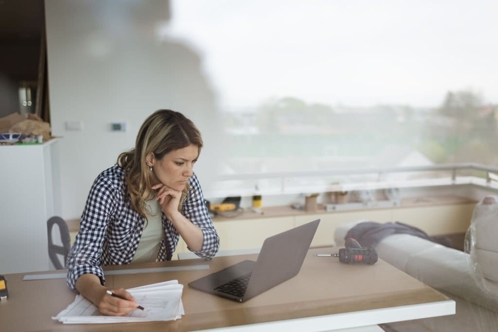 Professional woman sitting on a makeshift table in the middle of internal renovations happening in her household concentrating as to what is on her laptop screen whilst holding in her other hand a pencil to some architectural drawings