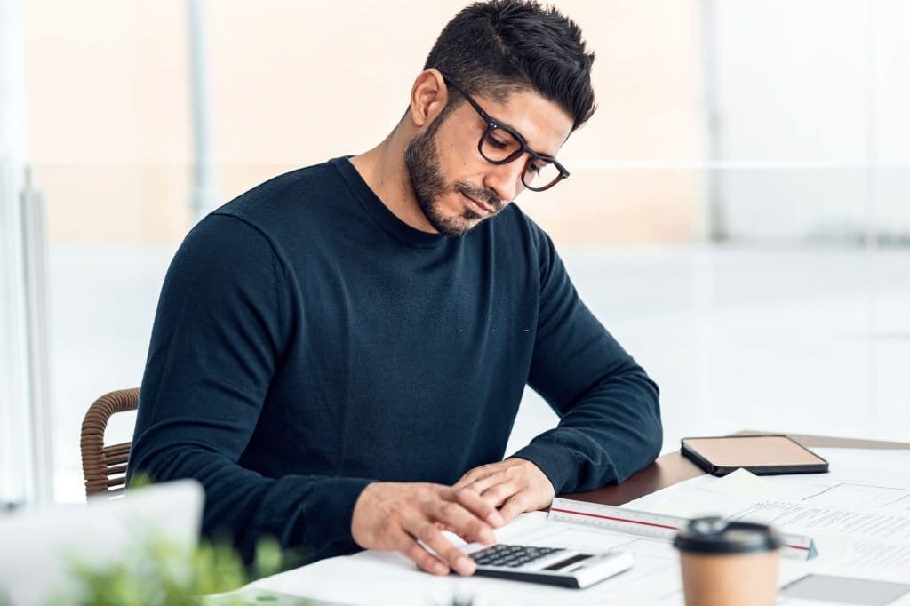 Young professional in dark brown rectangular glasses reviewing the costs implications on a silver and black calculator alongside all of the architect's drawings and planning consultants paperwork