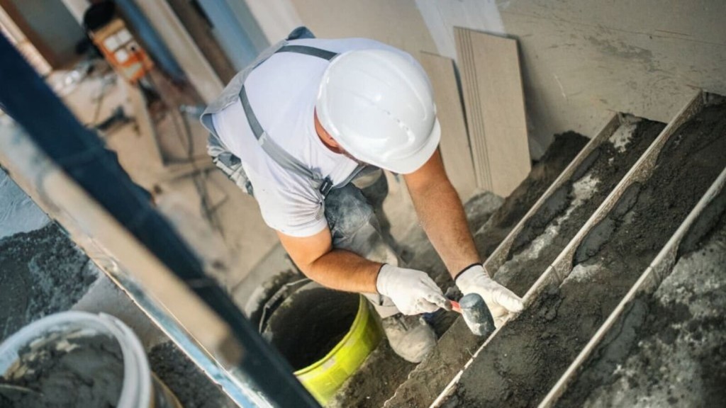 Construction worker in a white hard hat and gloves meticulously laying tiles on a new staircase, with a focus on precise alignment and spacing, in an indoor renovation site.