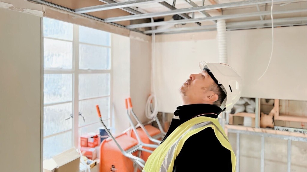 Electrician in a white hard hat and high-visibility vest inspecting overhead electrical wiring and metal conduits in a room under construction with natural light coming through the window.