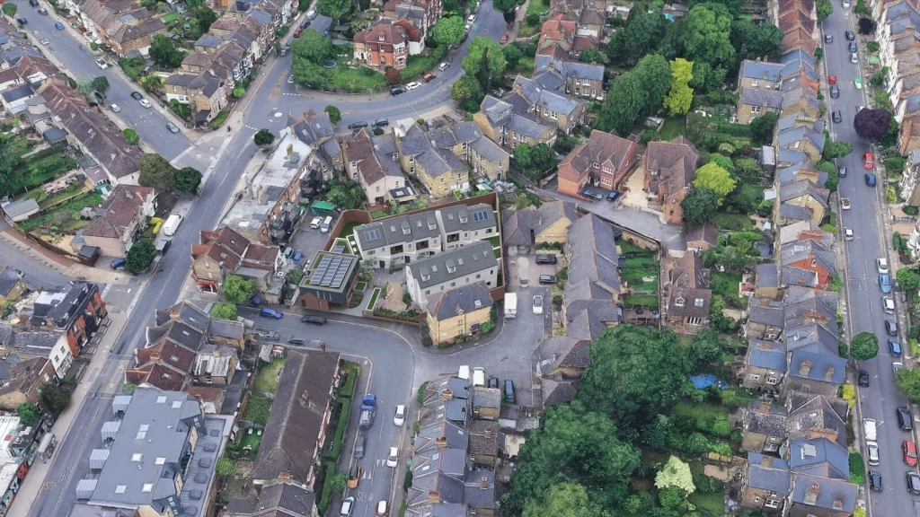 A Google Earth-style aerial view of London showing a mixed neighbourhood with terraced houses, small commercial plots and an inserted CGI scheme representing new-build homes and commercial units.