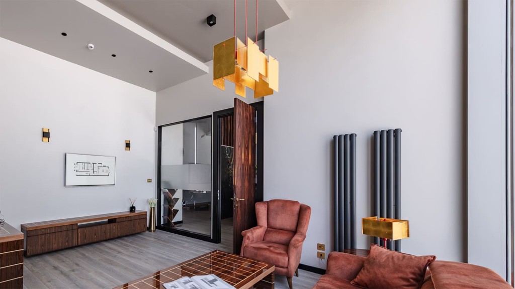 Chic office space with high ceilings, geometric hanging lights, brown suede armchair and sofa, and a wood media console against a backdrop of a modern glass partition and decorative wall fixtures.