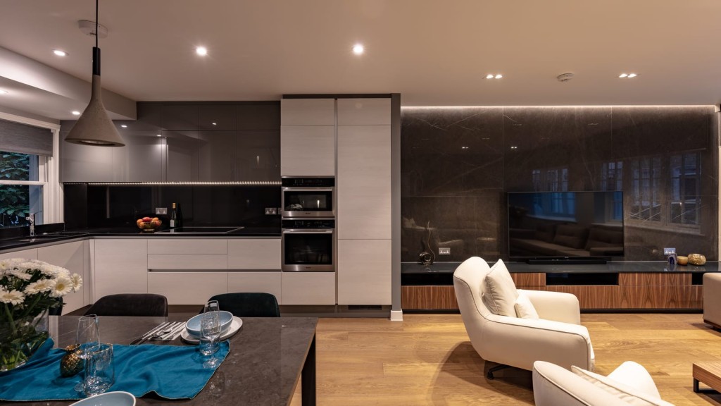 Sleek open-plan living space with a modern kitchen, featuring white cabinetry, integrated appliances, and a large dark backsplash, complemented by a dining area with elegant table setting and cosy lounge chairs.