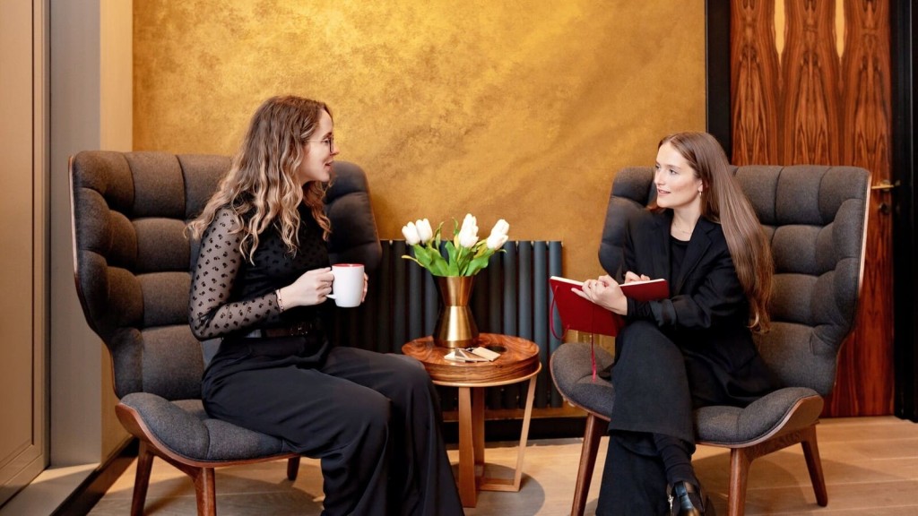 Two professional women engaged in conversation, seated on plush armchairs with a wooden side table and a vase of white tulips, against an elegant gold textured wall.