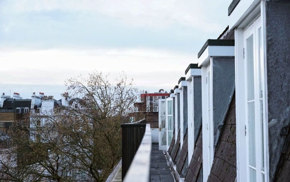 View along the sloped rooftops of a row of London houses with mansard extensions and white-framed dormer windows, showcasing urban residential architecture against a backdrop of tree canopies and a cloudy sky.