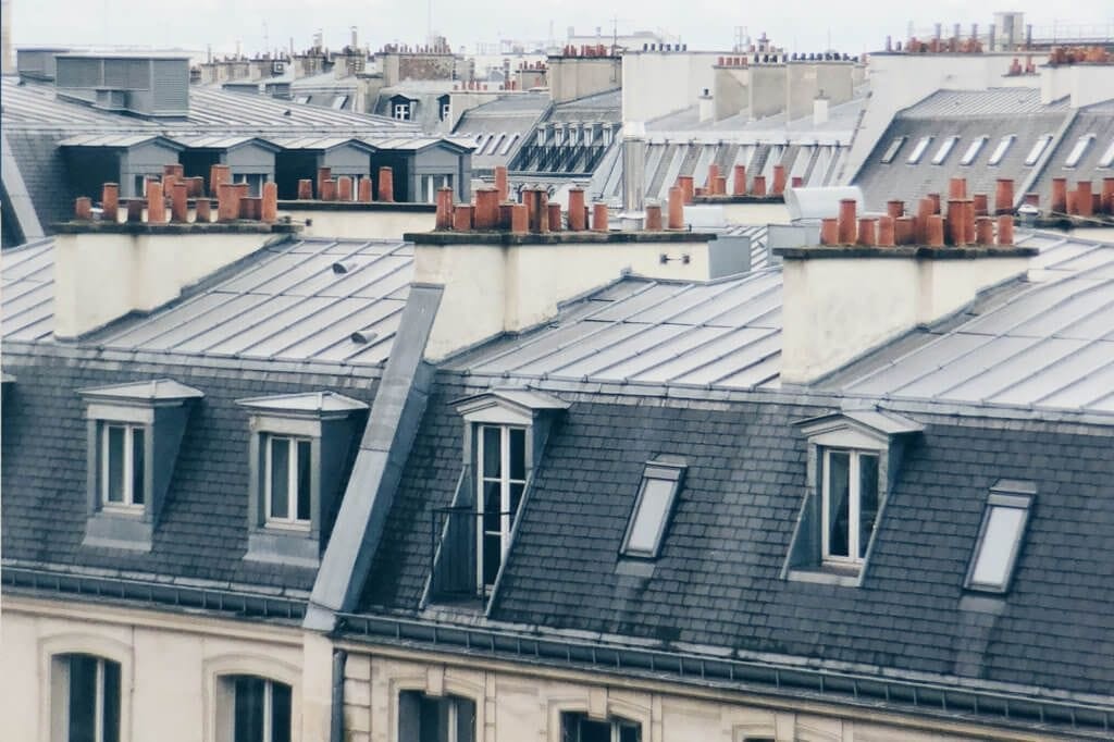 Close-up of a dense array of Parisian rooftops showing the classic gray zinc mansard roofs with dormer windows and red chimney pots, exemplifying traditional French urban architecture.