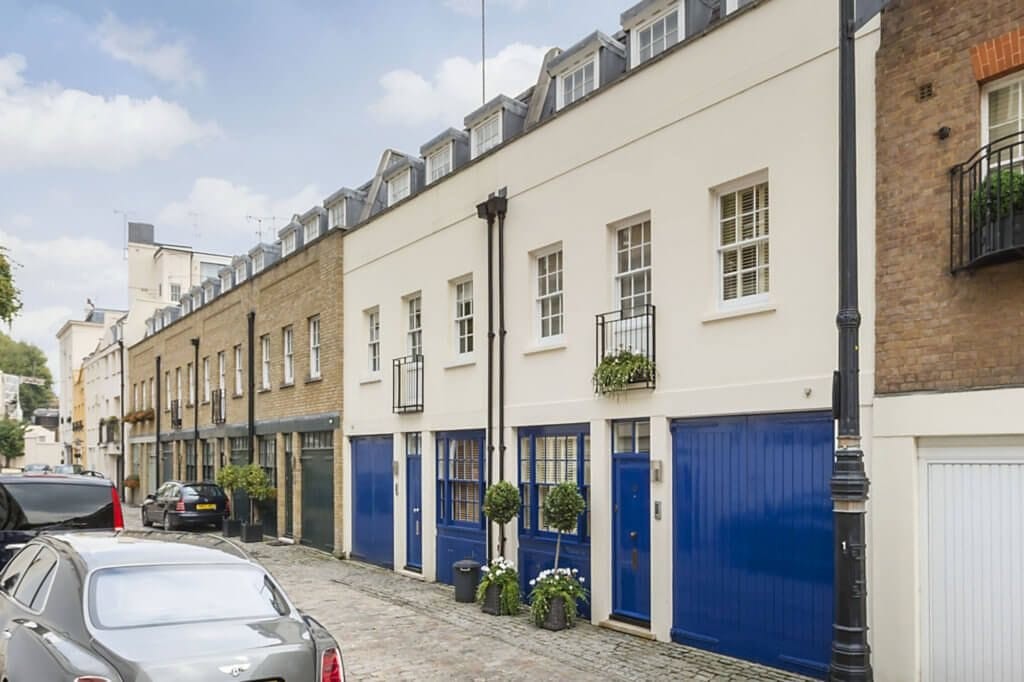 Charming mews houses in London with prominent mansard roofs featuring dormer windows, painted in neutral tones with contrasting blue garage doors, set on a cobblestone street.