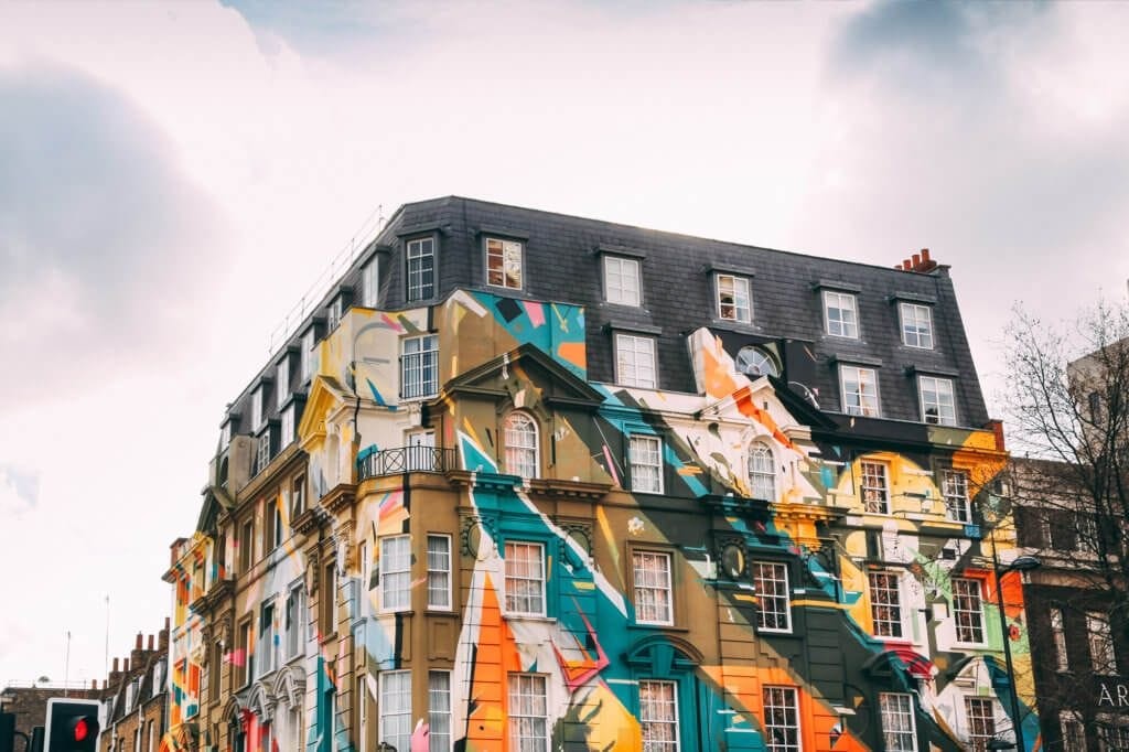Eye-catching multi-story building in London adorned with a vibrant, abstract mural over its classical architecture, featuring mansard roofing and wrought-iron balconies against a soft cloudy sky.