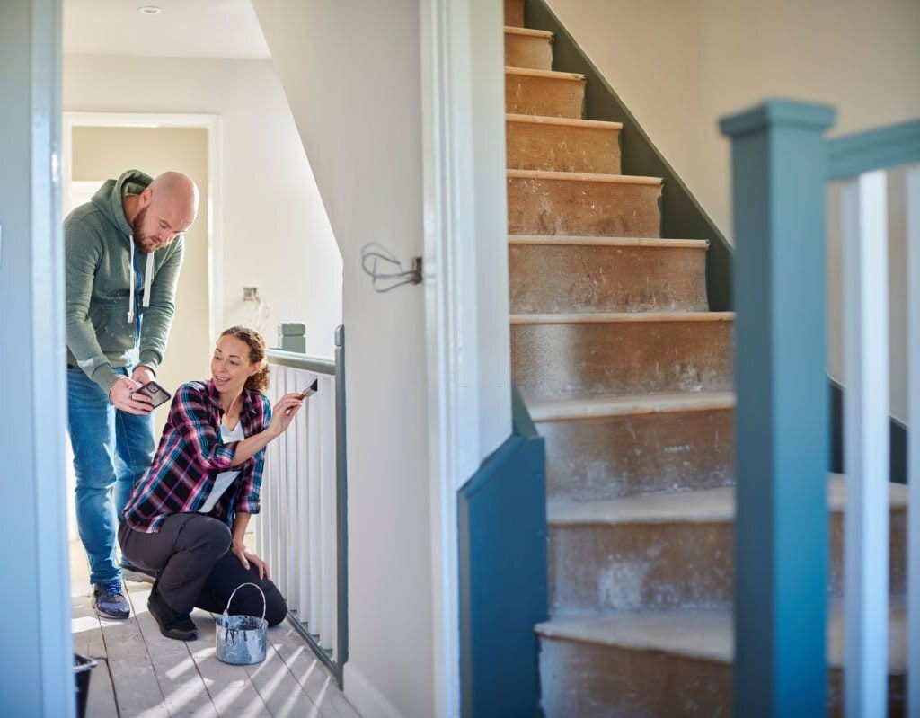 Interior painter crouching on her knees to paint the wooden stairs balustrade white whilst her colleague is standing on the side of her showing what further alterations can be done to the property